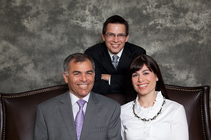 The image depicts three individuals posing together for a photograph two men stand behind a woman who is seated in front of them, all smiling at the camera.
