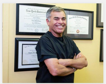 The image shows a man standing in front of framed diplomas, smiling at the camera. He has his arms crossed and appears to be posing for a formal portrait.